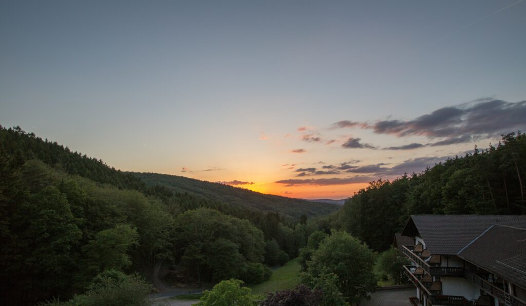 Sonnenuntergang über der bewaldeten Hügellandschaft des Naturparks Spessart mit traditionellem Gasthaus im Tal